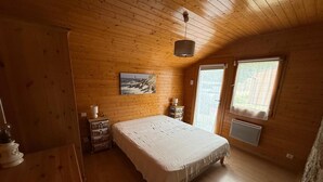 A wooden cabin bedroom with a bed, a nightstand, a lamp, a picture on the wall, and a window. - Charming Penty Breton, close to the beaches and town of Tregunc - 6 people max. (Trégunc)