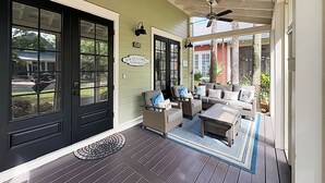 Un porche abrité avec un plancher en bois foncé, un coin salon avec un canapé et des fauteuils, et une porte noire ornée d’une pancarte de bienvenue.