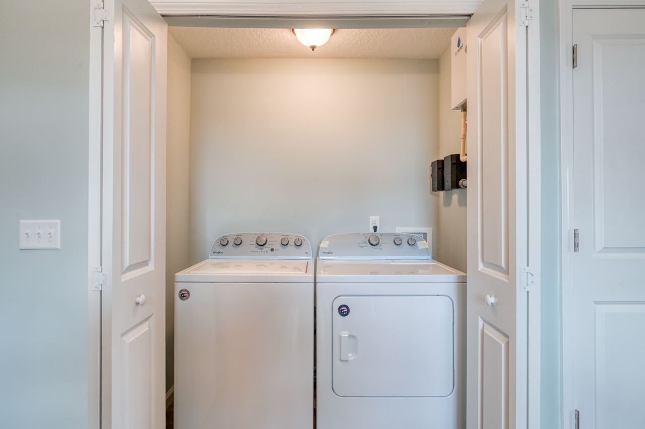 A laundry room with two stacked washers and dryers, a light fixture above, and a small shelf with a water heater.
