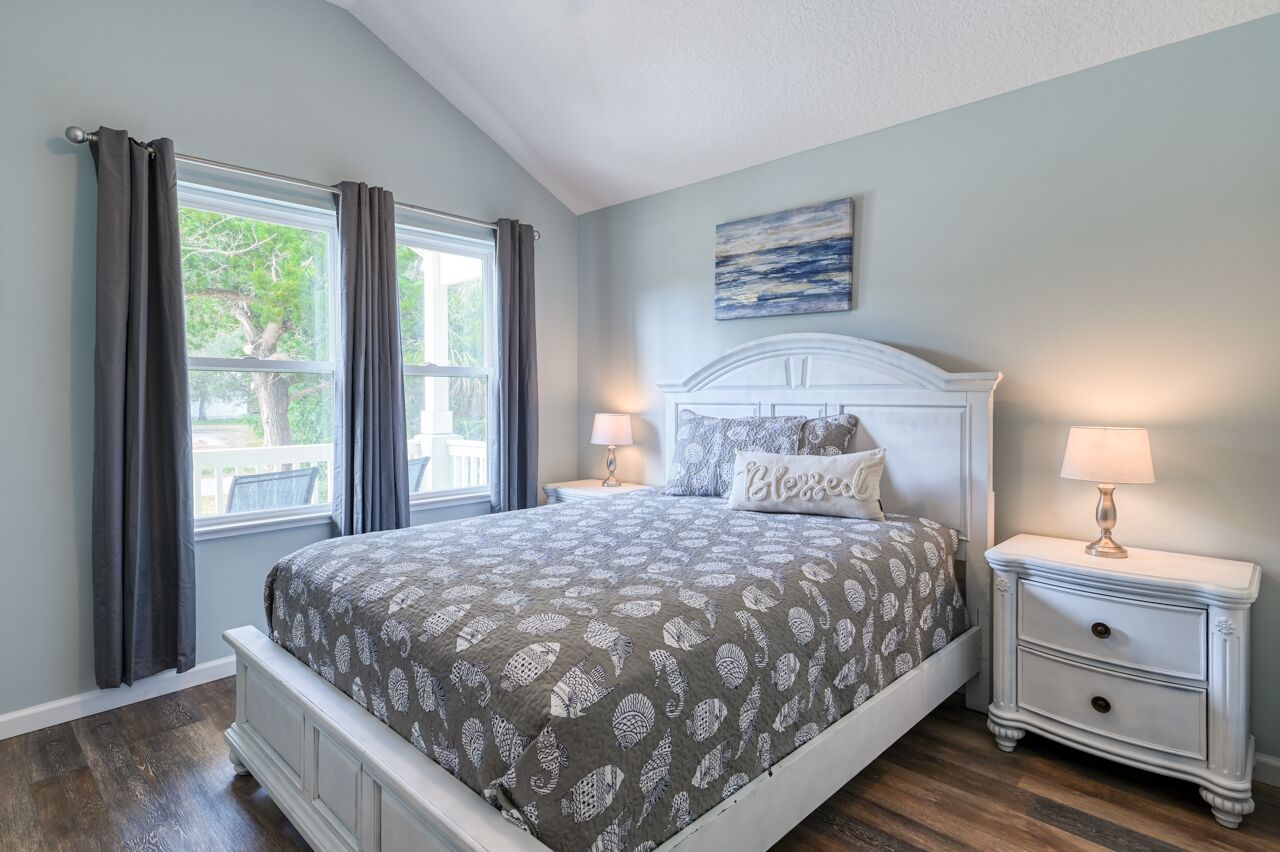 A bedroom with a bed, two bedside tables with lamps, a window with curtains, and a painting on the wall.