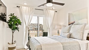 Une chambre à coucher avec un lit, des rideaux, un ventilateur de plafond, une plante en pot et une vue sur les maisons par la fenêtre.