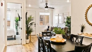Un coin repas avec une table en bois foncé, des chaises noires et un ventilateur de plafond. Des plantes en pot et un miroir ornent les murs.