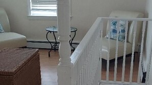 Una habitación amplia con techo alto, una escalera blanca, un ventanal con persianas, un reposapiés de mimbre y una silla blanca con cojín.