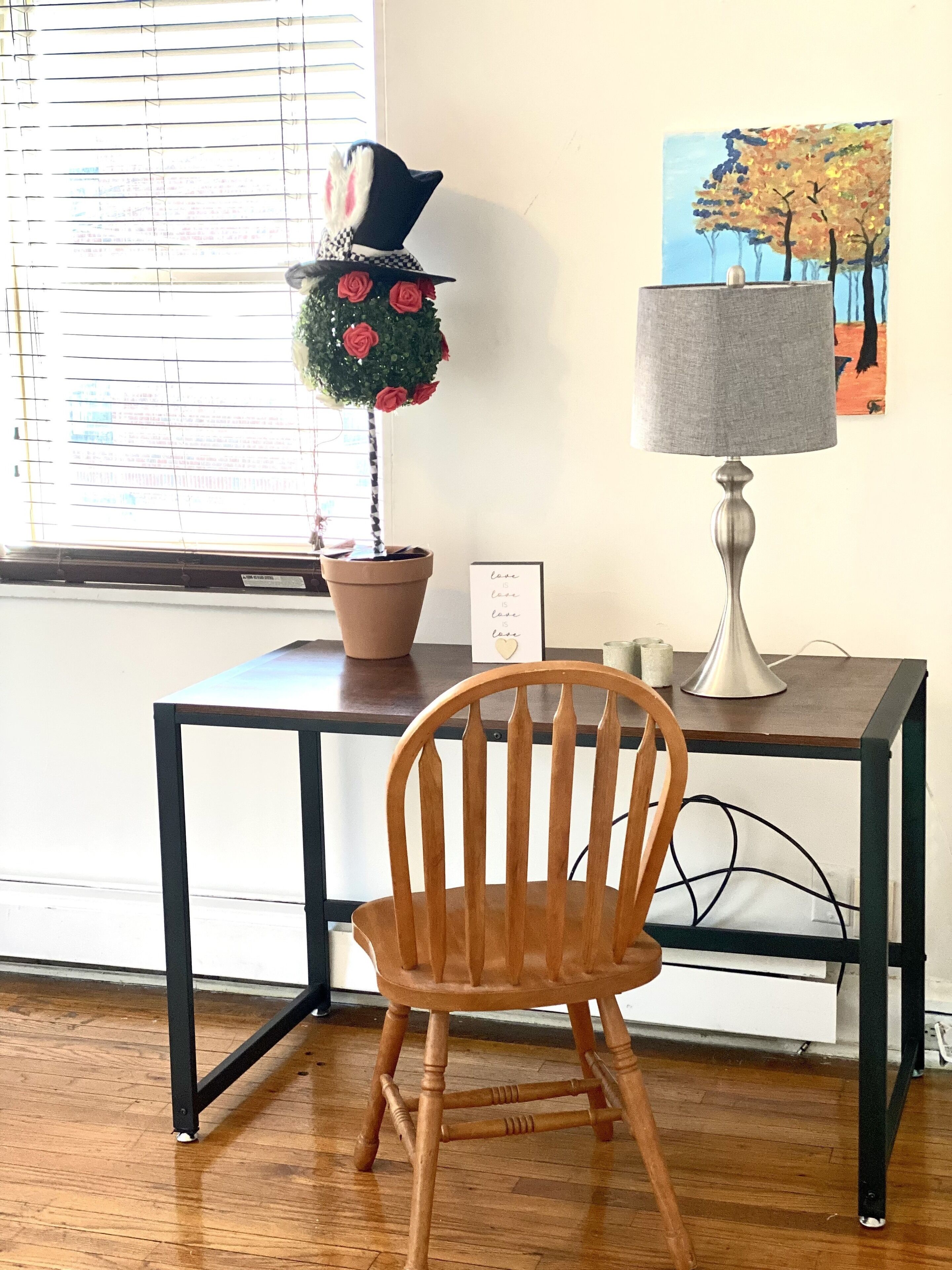 A wooden chair, a desk with a lamp, a potted plant with a topiary, and a painting on the wall.
