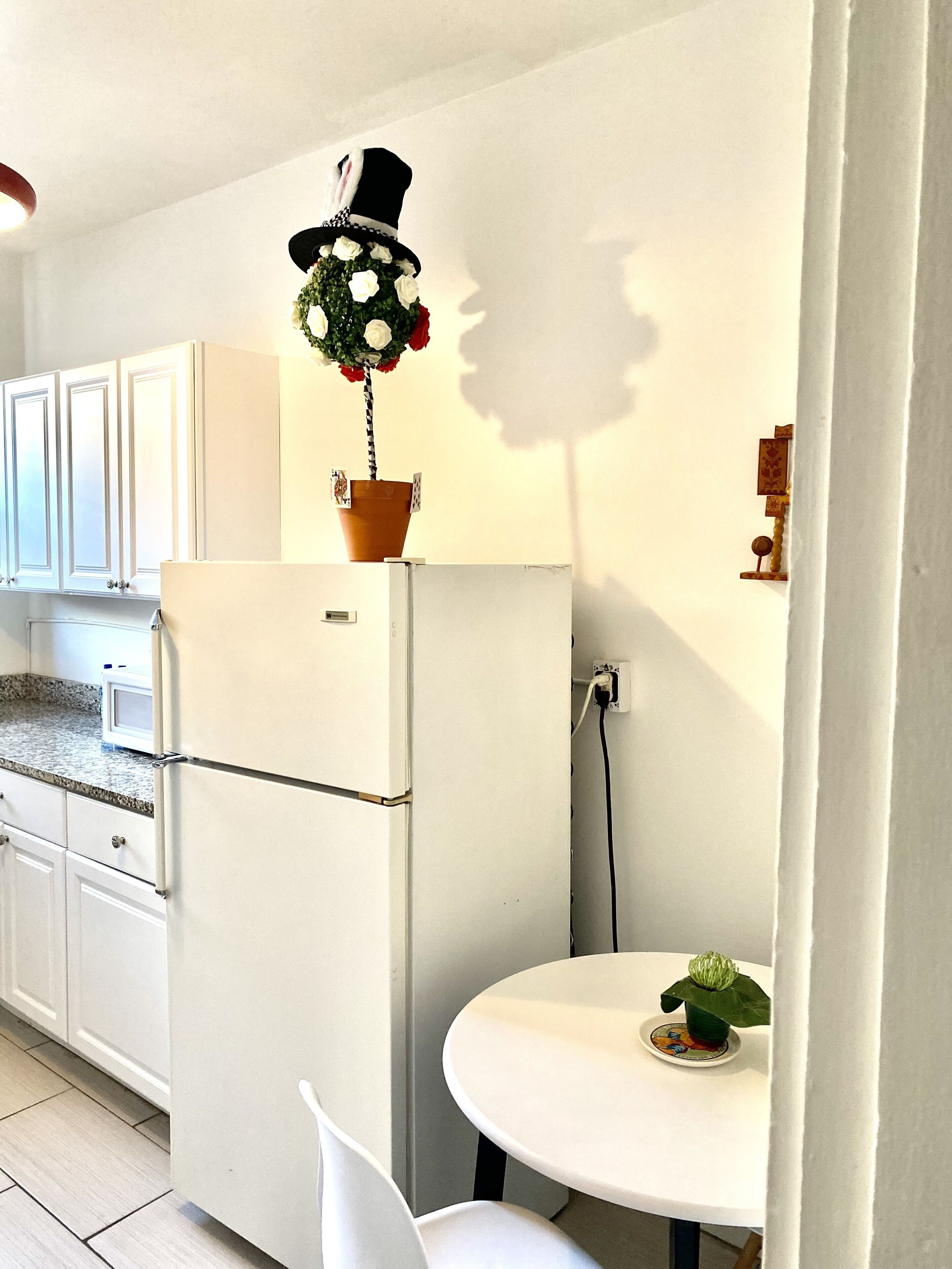 A compact kitchen with white cabinets, a white refrigerator, a small table with a decorative plant, and a shadow of a person on the wall.