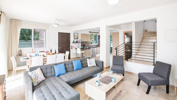 A modern living room with a grey sofa, a coffee table, and a grey armchair. There is a staircase leading to an upper floor. - Protaras Holiday Villa Kv60 (Pernera)