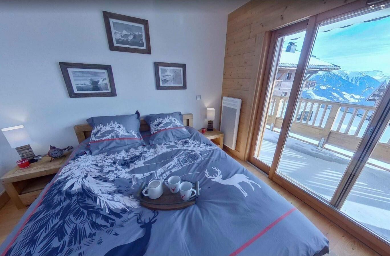 A bedroom with a large bed, a wooden headboard, a bedside table with a lamp, and a view of mountains through a large window.