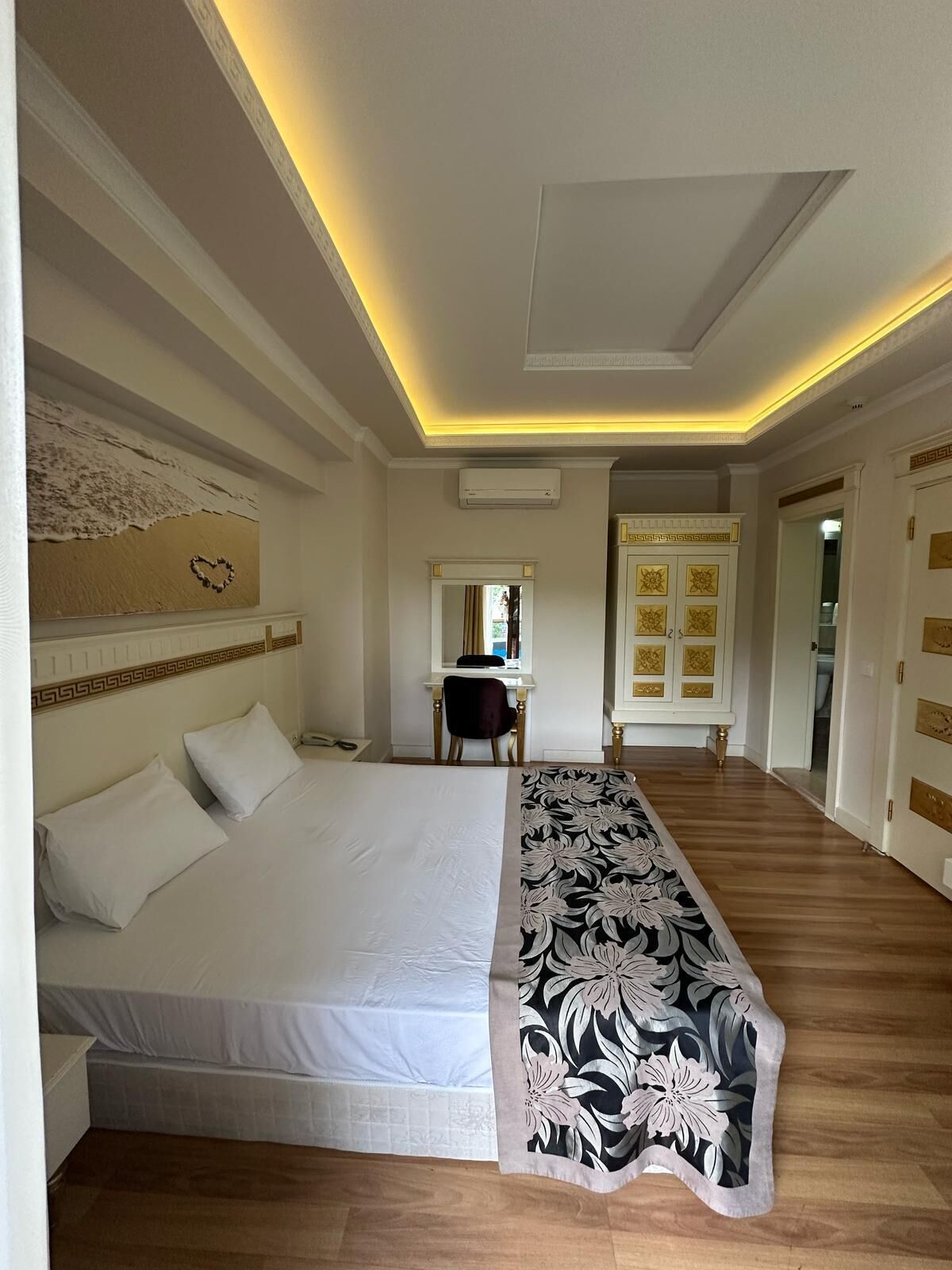 A hotel room with a large bed, a desk, and a chair. There is a mirror above the desk and a door with decorative panels.