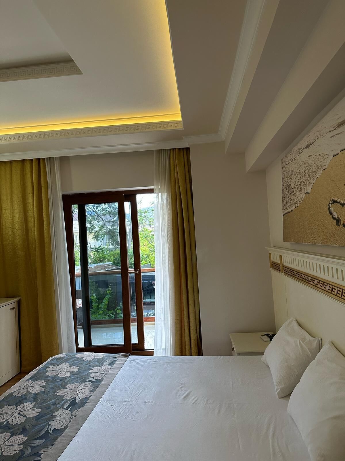 A hotel room with a bed, a nightstand, a lamp, and a view of greenery outside.