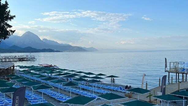A beach with rows of blue lounge chairs and green umbrellas, a pier extending into the water, and mountains in the background. - Barby Butik Otel (Kemer)
