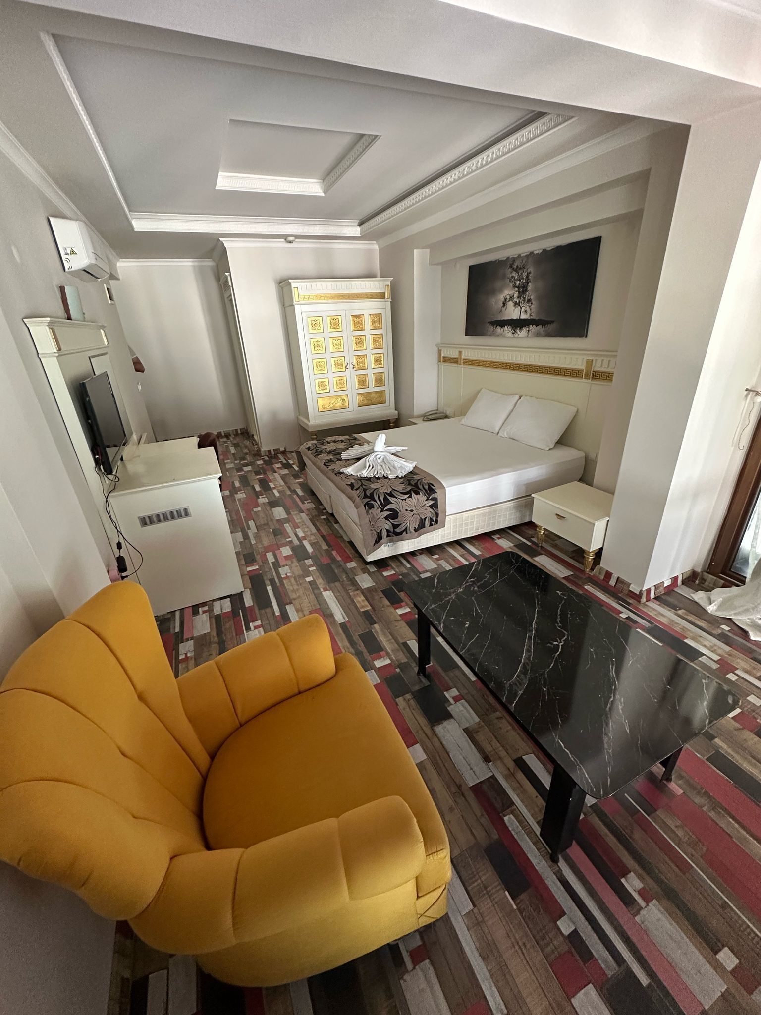 Habitación de hotel con cama, sofá, sillón amarillo, mesa de centro de mármol negro, alfombra estampada, armario blanco y un cuadro en la pared.