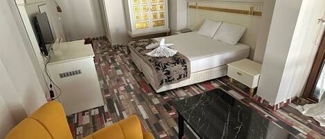 Habitación de hotel con cama, sofá, sillón amarillo, mesa de centro de mármol negro, alfombra estampada, armario blanco y un cuadro en la pared.