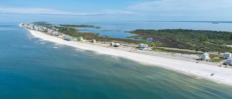 An aerial view of a coastal area with a long sandy beach, a small settlement with buildings, and a forested peninsula.