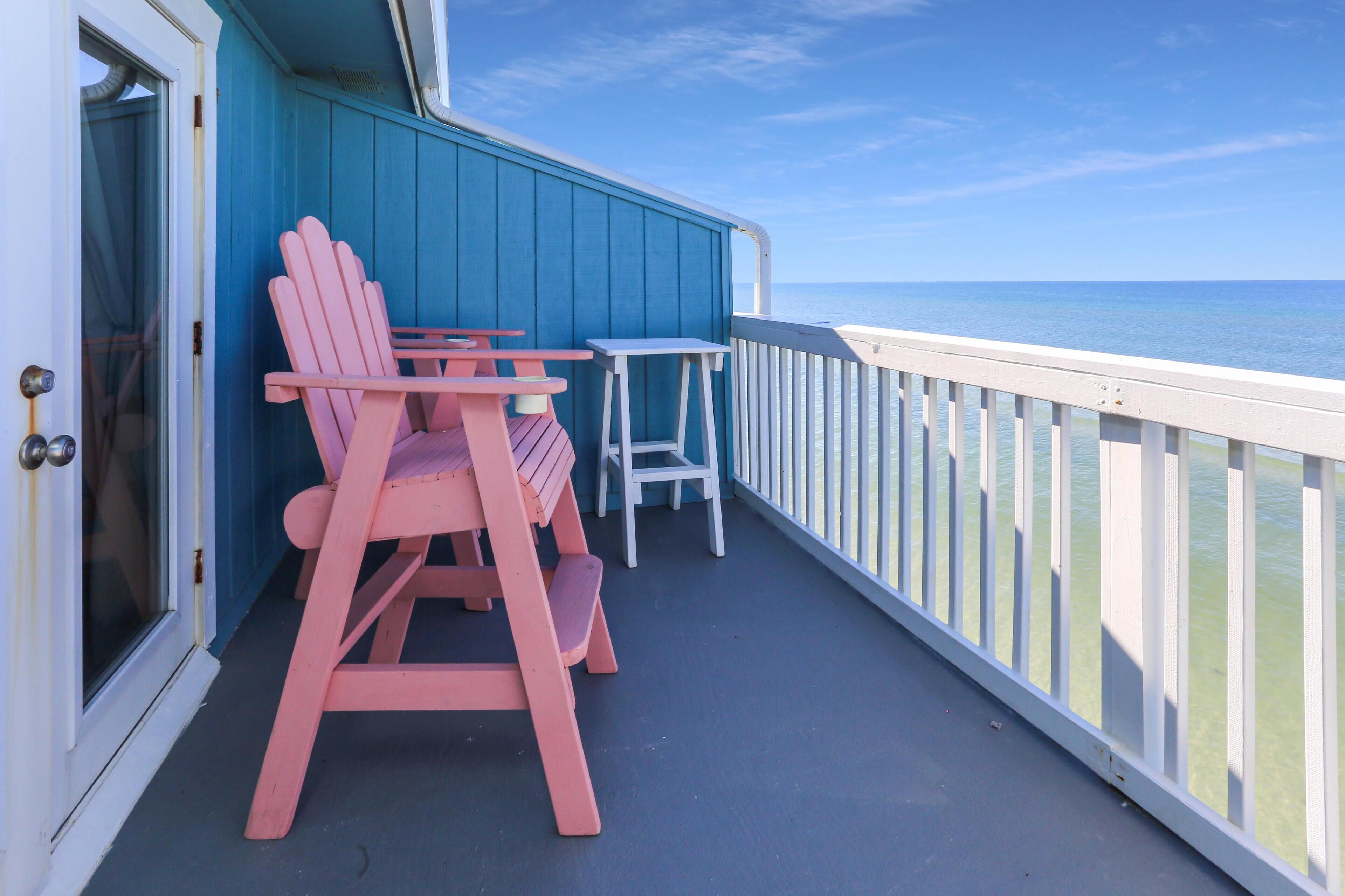 A balcony with a view of the ocean, featuring two pink Adirondack chairs and a small table.