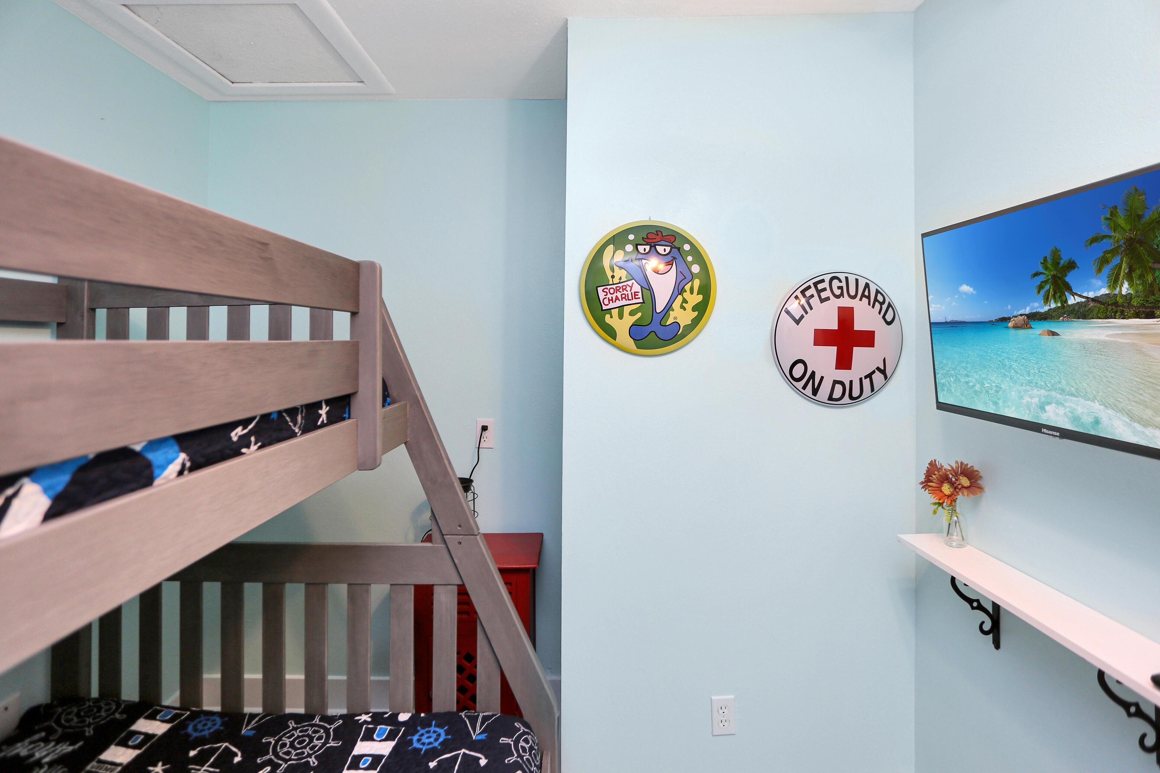 Un lit superposé avec une couvre-lit à motifs, un mur orné d’un panneau indiquant le poste de surveillance des sauveteurs, et un tableau encadré représentant une plage.