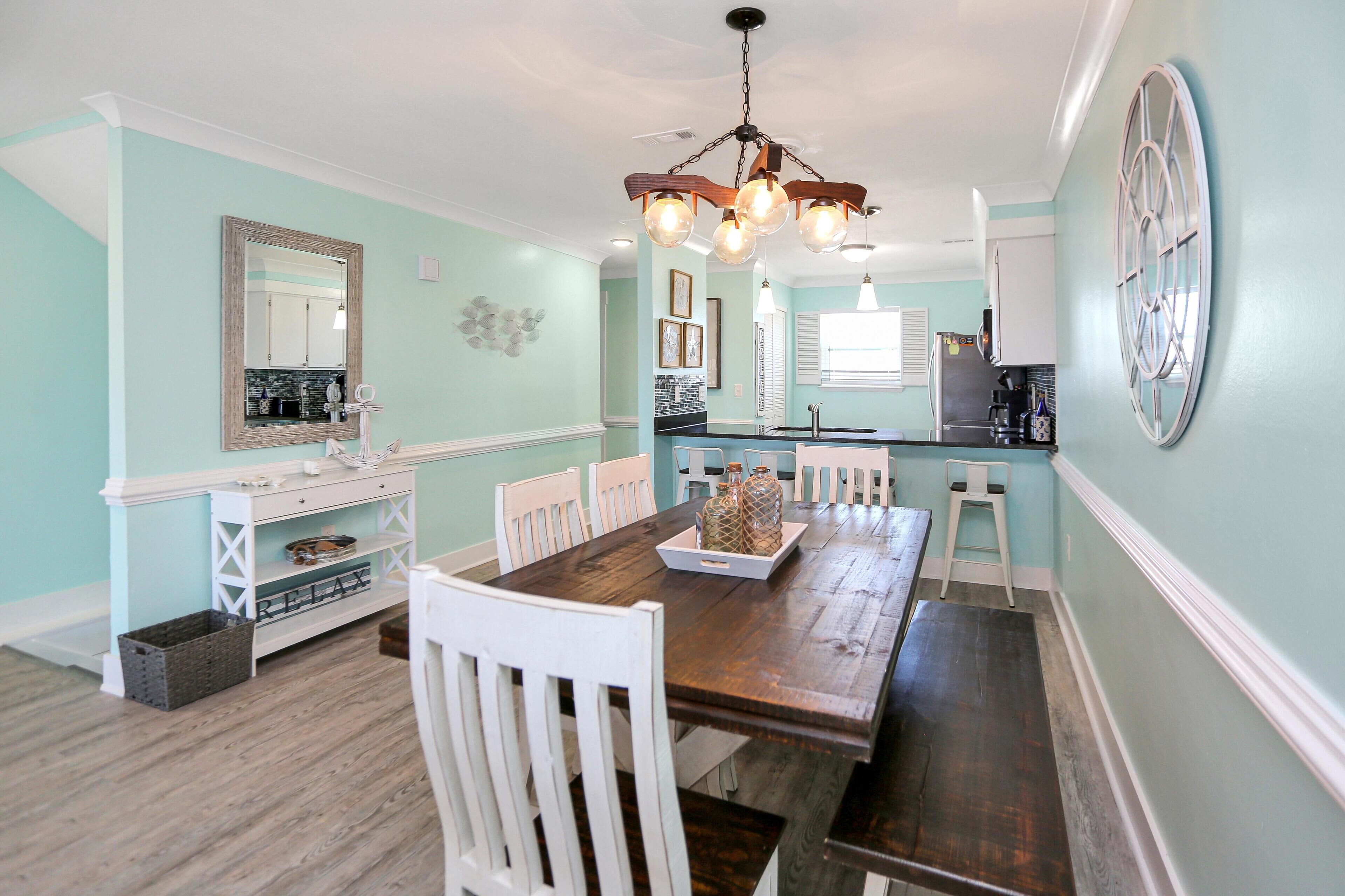 A dining area with a wooden table, white chairs, a chandelier, and a kitchen in the background.