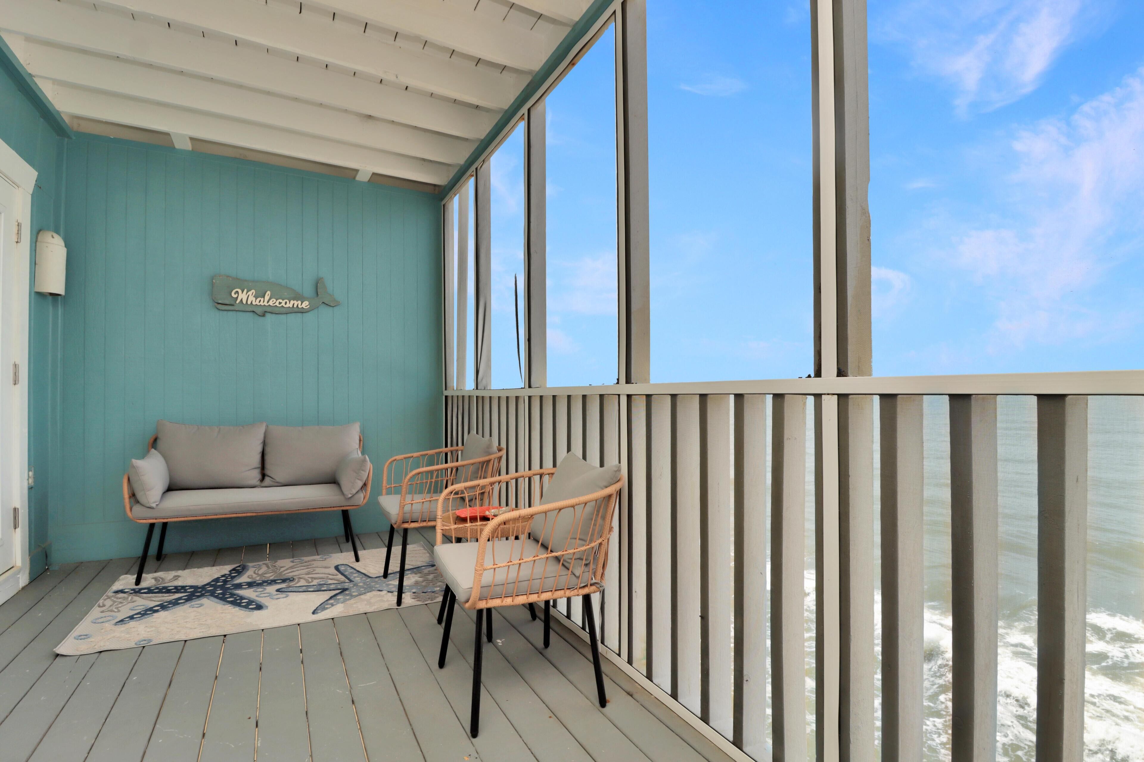 A balcony with a turquoise wall, a 'Whalecome' sign, a sofa, two wicker chairs, and a starfish rug.