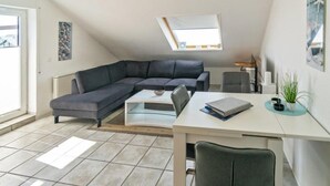Een moderne, compacte leefruimte met een grijze modulaire bank, een witte eettafel en een klein werkgedeelte. Een dakraam zorgt voor natuurlijk licht, en kunstwerken sieren de muren.