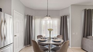 A dining area with a round table, chairs, a hanging light fixture, and a view through a window with curtains. - Luxury Pool Villa, sleeps 14, 5 mins to Disney, Games Room, gated resort (Kissimmee)
