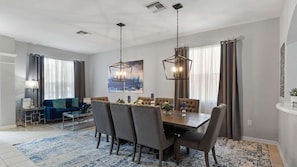Uma sala de jantar com uma mesa posta para seis pessoas, rodeada por cadeiras, e um sofá ao fundo.