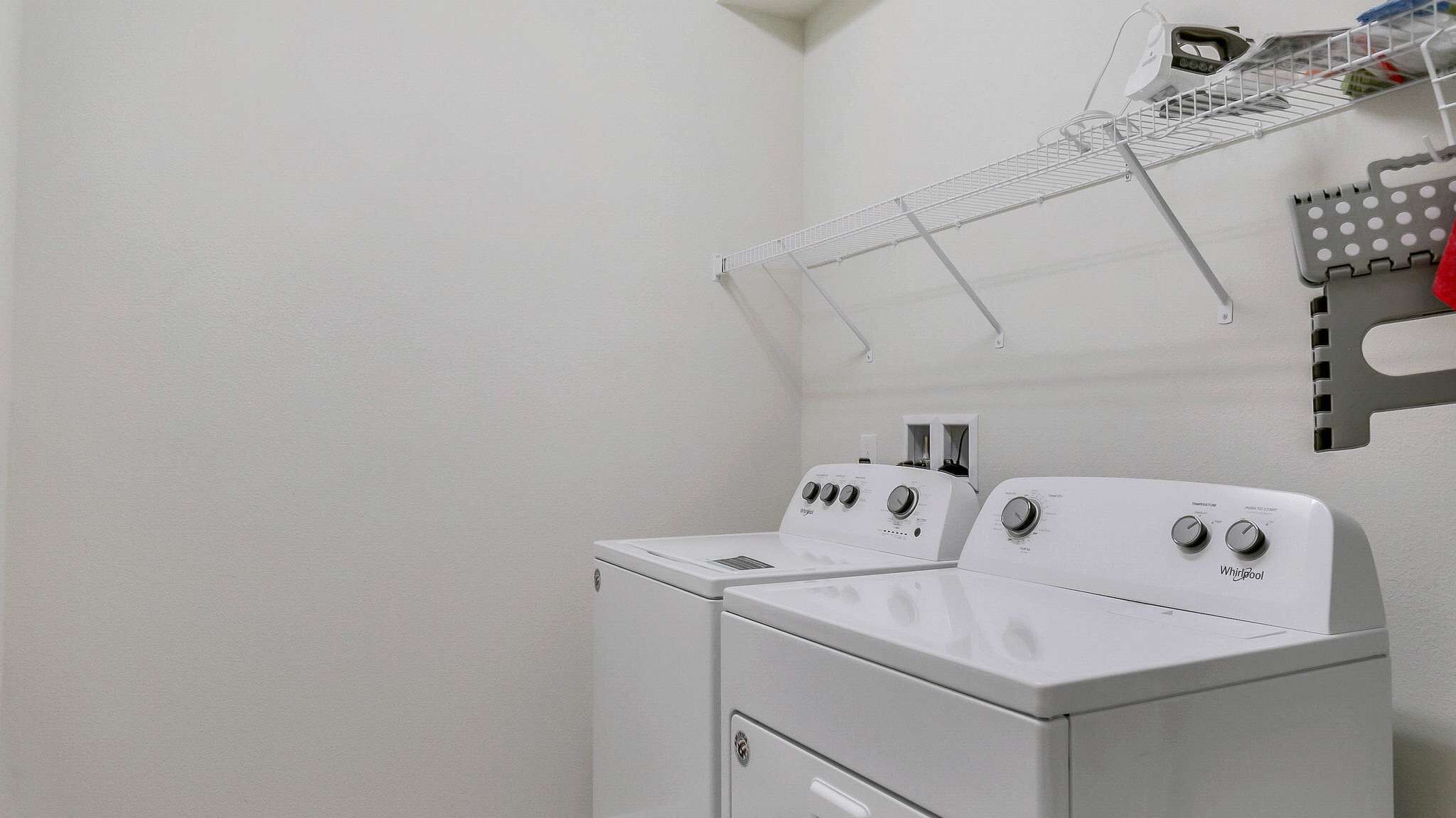 A laundry room with two white Whirlpool washers and dryers, a metal drying rack, and a white wall.