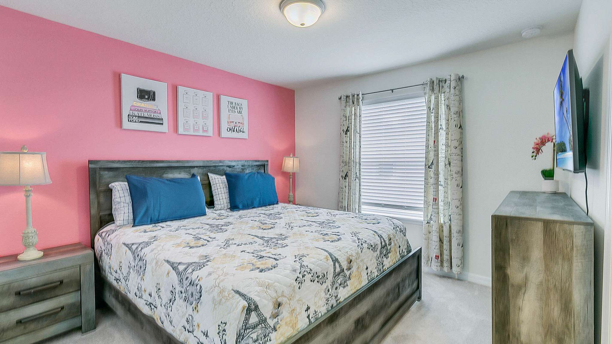 A bedroom with a pink wall, a bed with blue pillows, a nightstand with a lamp, and a TV on a wooden stand.