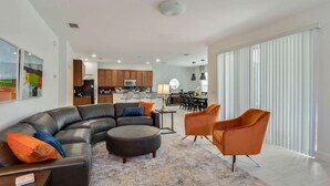 Un salon moderne avec un canapé d’angle, un pouf et deux fauteuils. On aperçoit une cuisine et un coin repas à l’arrière-plan.