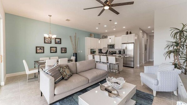 A modern living room with a sofa, coffee table, and chairs. There is a dining area with a table and chairs. The kitchen is visible with cabinets and appliances. - Resort Home, private pool, gated community, great location! (Davenport)