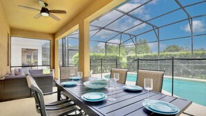 A sunlit dining area with a pool view, featuring a table set with blue plates and glasses, wicker furniture, and a ceiling fan.