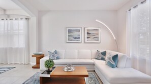 Una sala de estar moderna con un sofá modular blanco, dos cuadros abstractos enmarcados, una mesa de centro de madera con una planta y una lámpara de pie.