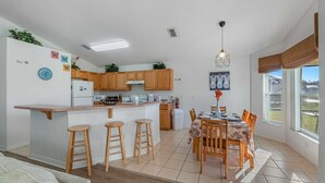 Una cocina moderna con armarios de madera, una isla central con taburetes, un comedor con mesa y sillas, y vistas al exterior a través de amplios ventanales.