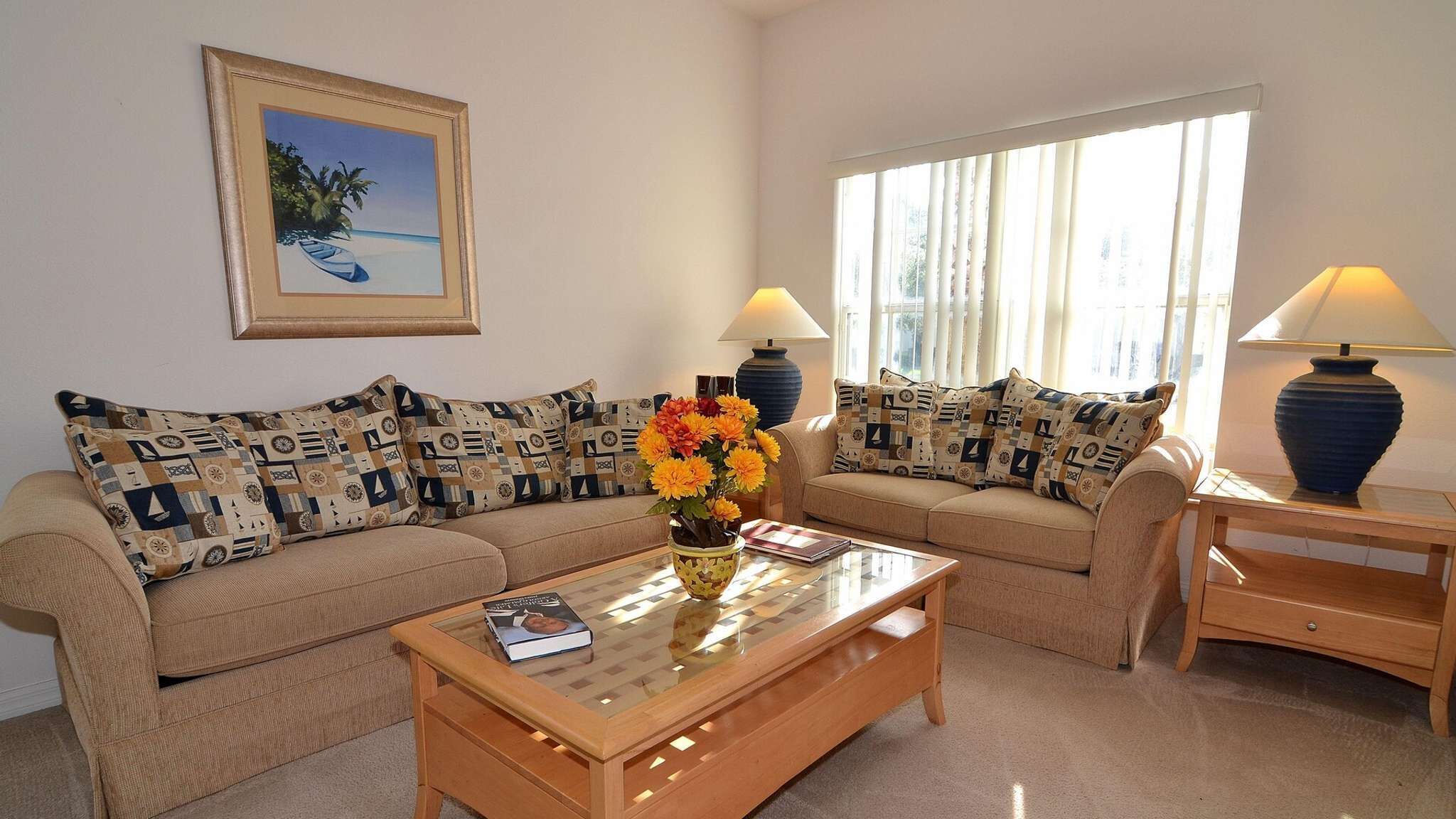 A living room with a beige sofa set, patterned cushions, a glass coffee table, a vase with flowers, a framed picture on the wall, and two lamps.