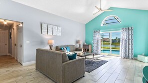A living room with a grey sofa, a coffee table, and a lamp. There is a pool visible through the glass doors.