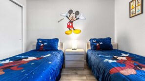 A room with two beds, each with a Mickey Mouse-themed duvet. There is a nightstand with a lamp between the beds.
