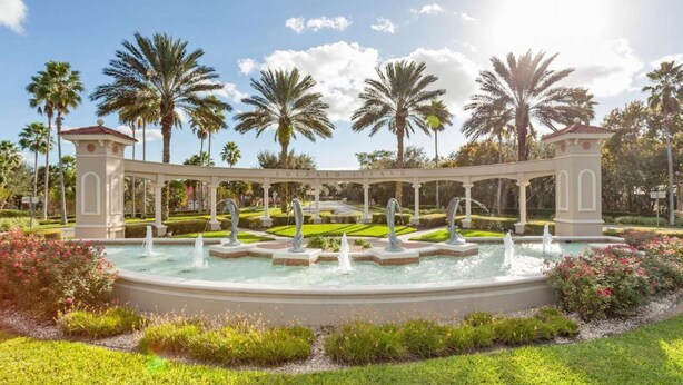 A circular fountain with multiple water jets, surrounded by a white pavilion with columns, palm trees, and well-manicured gardens.