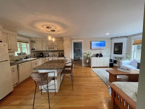 A modern kitchen with a central island, wooden flooring, and white cabinetry. There is a dining area with a wooden table and chairs, and a living room with a sofa and a television.