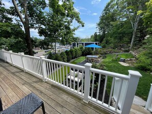 A wooden deck with a white railing, overlooking a well-maintained garden with a patio area, a blue umbrella, and a stone wall. - Hot Tub, Putting Green, 5 min walk to the beach, shops & restaurants. (Ogunquit)