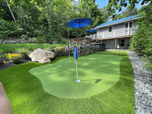 A well-maintained artificial green golf course with a flagstick, surrounded by a stone wall, a large rock, and a house with a blue roof in the background.