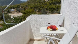 A balcony with a table set for two, featuring a plate of fruit and a glass of red wine. The balcony overlooks a mountainous landscape with greenery and a few buildings in the distance. - Golden Star Praxitelous (Aegina)