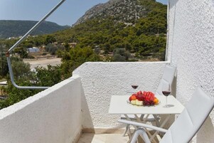 Un balcón con una mesa dispuesta para dos, mostrando un plato de fruta y una copa de vino tinto. El balcón da a un paisaje montañoso con vegetación y algunos edificios a lo lejos.