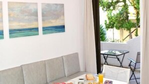 A dining area with a white table set for breakfast, including fruits, pastries, and beverages. There are three paintings on the wall depicting a beach scene. - Golden Star Praxitelous (Aegina)
