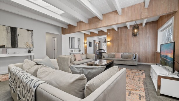 A spacious living room with a large sectional sofa, a coffee table, and a TV on a wooden stand. There is a large abstract painting on the wall and a rug on the floor.