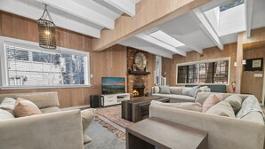 A modern living room with a fireplace, large windows, and a high ceiling with exposed beams.