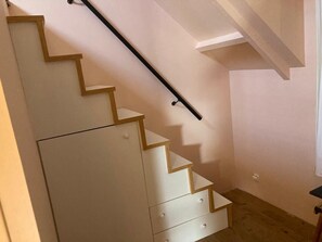 Una escalera con peldaños de madera y barandilla negra, que lleva a un rellano con pared blanca y un pequeño armario blanco.