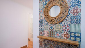 A hallway with a tiled wall featuring a variety of geometric and floral patterns. A wooden shelf is mounted on the wall, and a round mirror with a woven frame is centered above it. - Bella Portet - cosy apartment in El Portet (Moraira)