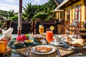 A breakfast table with various dishes, a drink, and a tropical outdoor setting. - Pousada Pedras Secas (Fernando de Noronha)