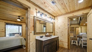A wooden cabin interior with a large mirror above a dark wooden vanity, a bed with a window, and a staircase leading to an upper level. - Large River front cabin overlooking Glover River (Broken Bow)