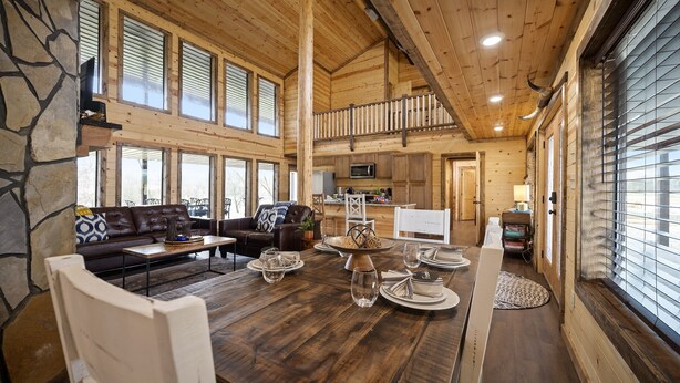A spacious wooden interior with a dining area, a living room with a sofa and coffee table, and a kitchen area. - Large River front cabin overlooking Glover River (Broken Bow)