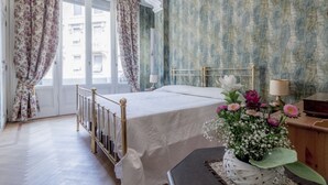 Une chambre à coucher avec un lit double, des rideaux à motifs floraux, un papier peint à dessins et une table de chevet avec une lampe et des fleurs.