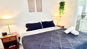 A neatly made bed with a dark blue quilt, two bedside lamps, a small wooden nightstand with a book, and a hanging plant. - Southwinds #9 for 3, King Studio Steps to Beach (Hollywood)